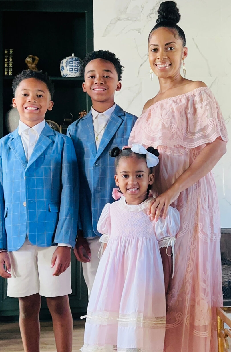 A woman and three children dressed formally stand in a living room, smiling at the camera. The boys wear blue blazers; the girl and woman wear light pink dresses. Ebony Boyd’s keen musical ear and appreciation for visual art shine through in their coordinated, elegant attire and poised demeanor.