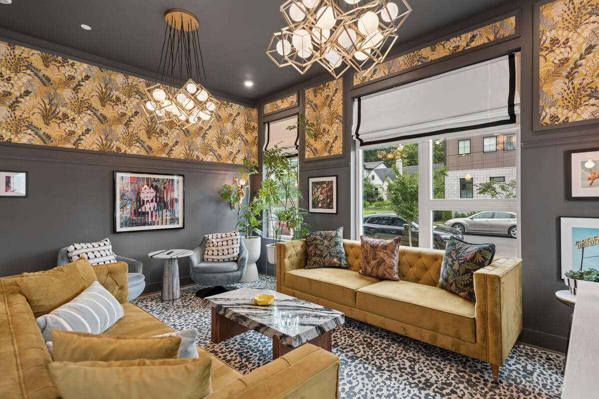 Modern living room in Unit 101 at 1310 Lischey Ave, Nashville, featuring mustard yellow sofas, patterned wallpaper, geometric lighting fixtures, and large windows showing a street view outside. Artwork and plants add decorative touches.