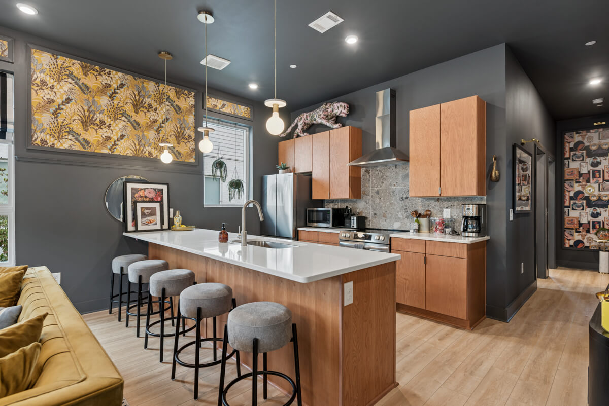 Modern kitchen at 1310 Lischey Ave, Unit 101, Nashville, featuring a large island, wooden cabinets, stainless steel appliances, patterned wallpaper, and four cushioned bar stools under pendant lights.