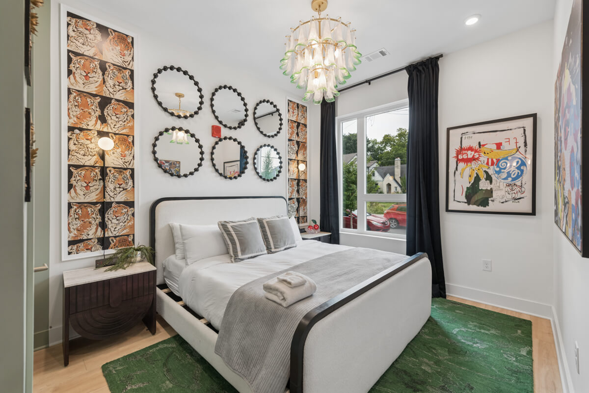 Modern bedroom in Unit 101 at 1310 Lischey Ave, Nashville, featuring a cozy bed, green rug, bedside table, round mirrors on the wall, a large window with dark curtains, and eclectic wall art.
