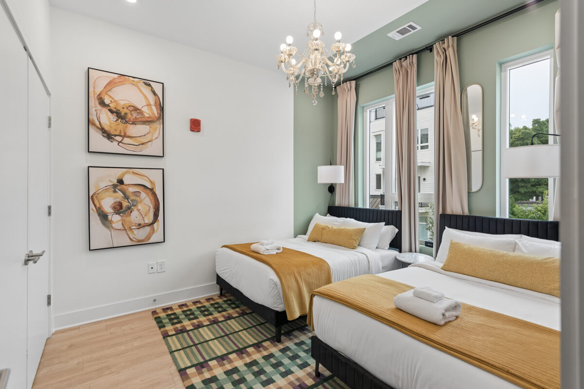 Modern bedroom at 1310 Lischey Ave, Unit 101, Nashville, featuring two twin beds with yellow blankets, folded towels, a chandelier, abstract wall art, large windows with curtains, and a checkered rug.