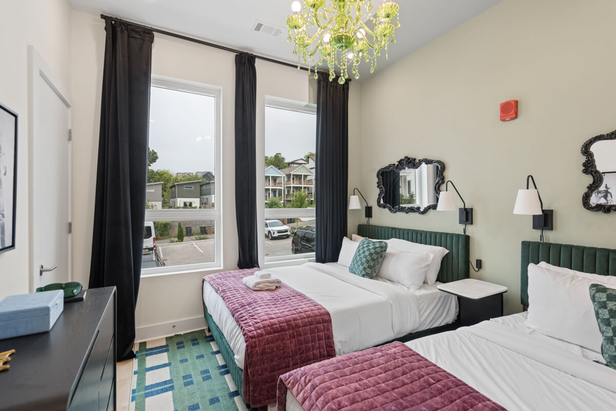 Modern bedroom at 1310 Lischey Ave, Unit 101, Nashville, featuring two double beds with purple throws and green velvet headboards. Enjoy large windows with black curtains, a striking green chandelier, stylish wall art, and an exterior view of parked cars and buildings.