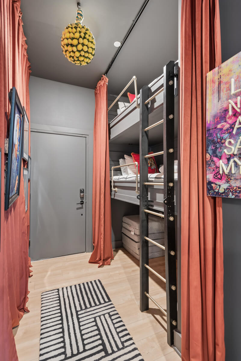 A narrow room at 1310 Lischey Ave, Unit 101 in Nashville features bunk beds, orange curtains, a black ladder, a geometric rug, wall art, and a closed gray door.