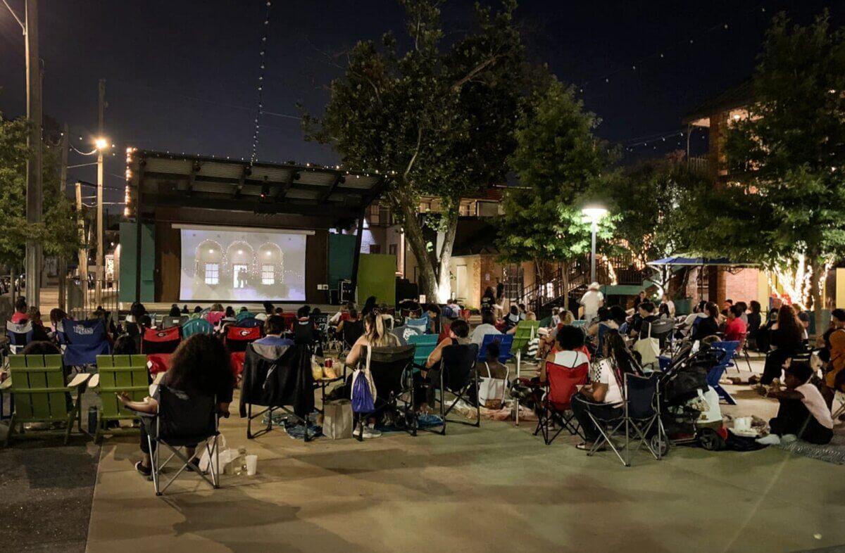 Where to Watch Spooky Movies Under the Stars Around Memphis - 2 People sit in lawn chairs watching a movie projected on an outdoor screen at night in a public plaza with trees and string lights.