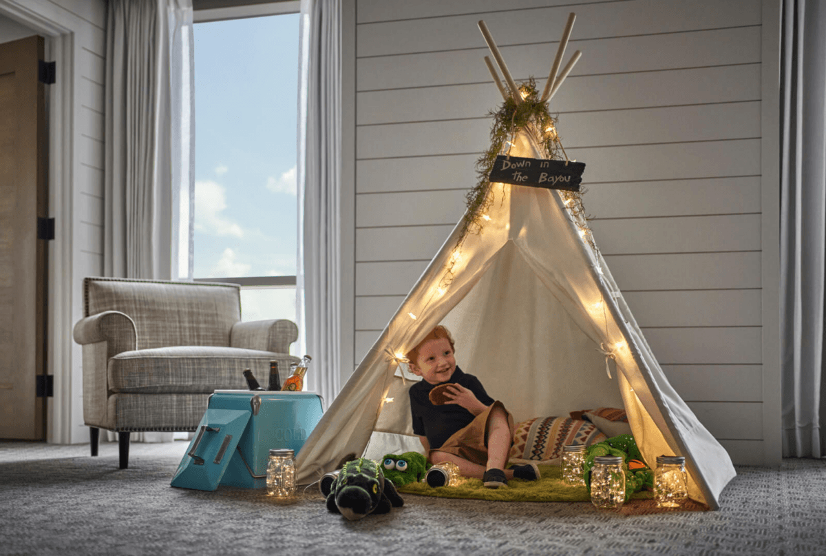 A young boy sits inside a small indoor teepee decorated with lights and greenery, holding a stuffed animal, with toys and pillows around him in a cozy, carpeted room—one of the unique hotel features designed to make families feel right at home.