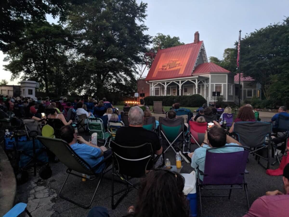 Where to Watch Spooky Movies Under the Stars Around Memphis - 3 A group of people seated in lawn chairs outdoors faces a small stage with lights, set in front of a house with a red roof during the evening.