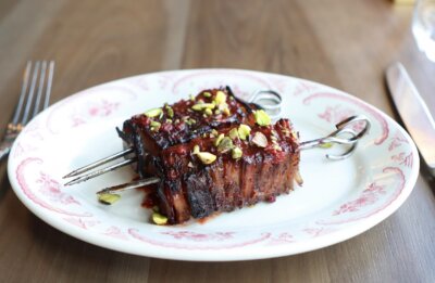 Two skewers of grilled meat topped with chopped pistachios on a decorative white plate, with a fork and knife on a wooden table.