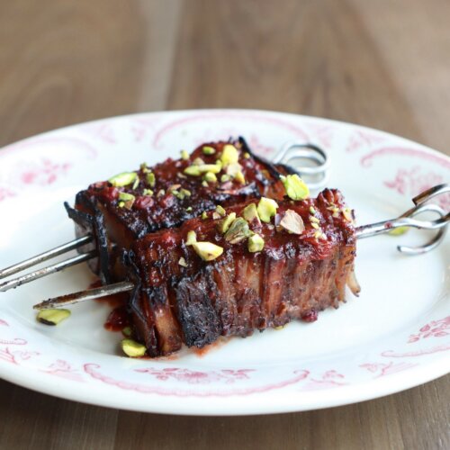 Your New Go-To Skewers for Tailgate Season - 2 Two skewers of grilled meat topped with chopped pistachios on a decorative white plate, with a fork and knife on a wooden table.