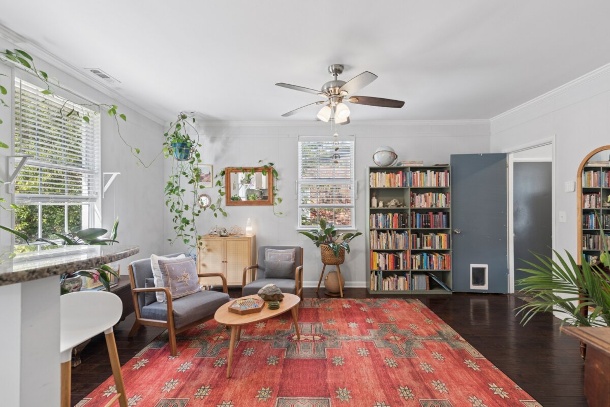 A living room in a Nashville home with a red patterned rug, two grey armchairs, a wooden coffee table, a tall bookshelf, and various potted plants.