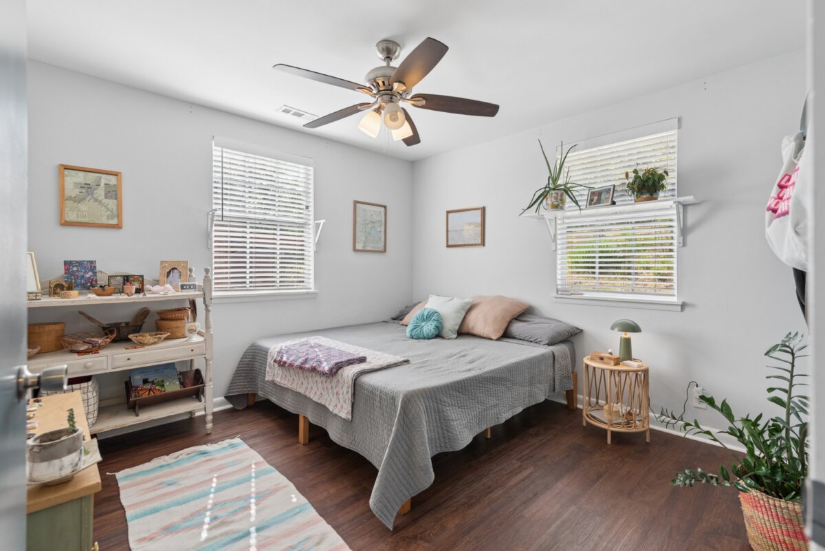 A bedroom in a Nashville home featuring a grey-quilted bed, a ceiling fan, a white shelving unit, and two windows with blinds.