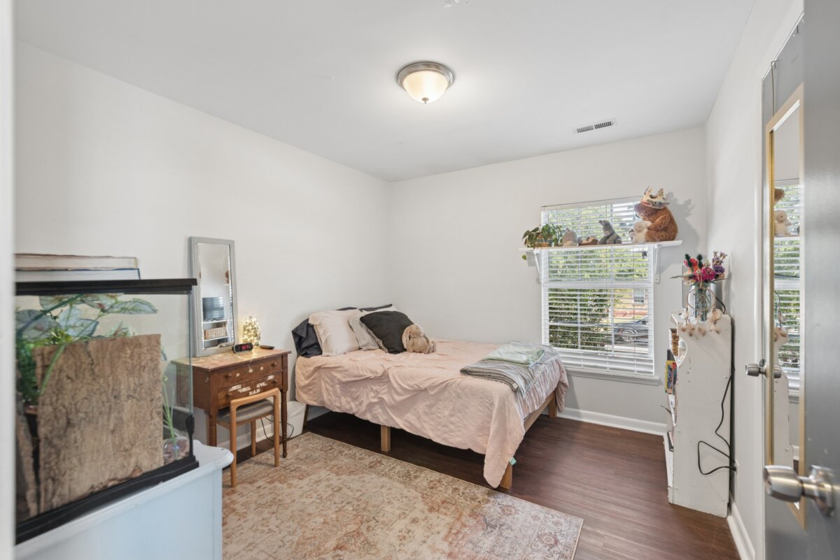 A bedroom at this Nashville home features a bed with a pink comforter, a wood vanity, a glass terrarium, and a window shelf holding stuffed animals and plants.