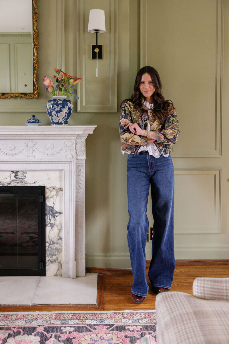 From ICU Nurse to Interior Designer: Meet Gretchen Rose of Kentucky Rose Designs - 2 A woman with long brown hair stands with arms crossed in front of a fireplace, wearing a floral jacket over a white blouse and dark blue wide-leg jeans.
