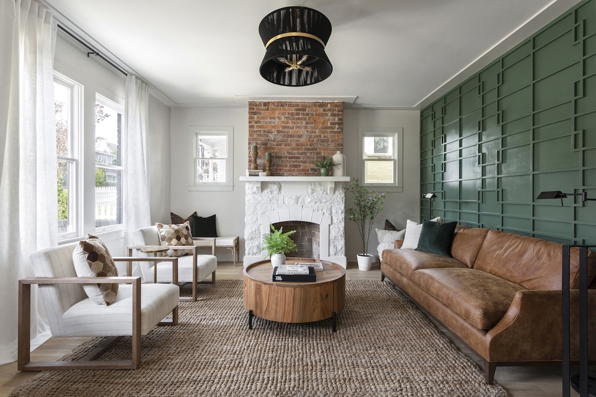 Living room with a brown leather sofa, two white armchairs, a round wooden coffee table, and a white stone fireplace with a brick chimney breast against a green paneled wall.