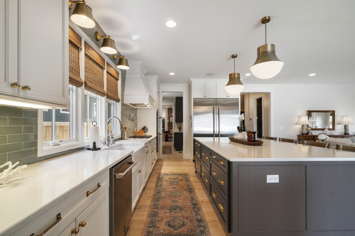 A white kitchen featuring a large central island with dark grey cabinets, white countertops, brass hardware, and a patterned runner rug on the wood floor.