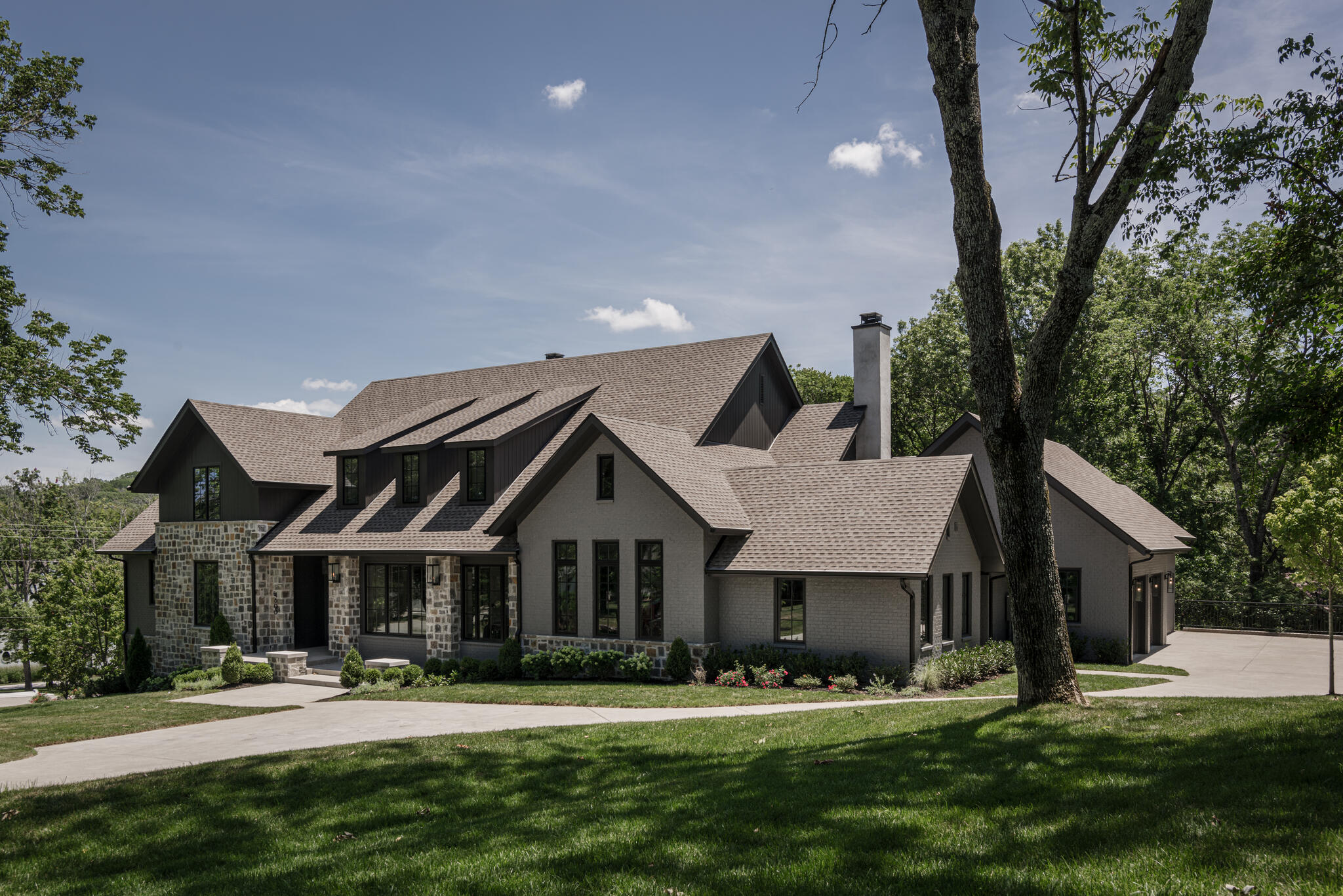 A large, multi-level suburban house with a gray brick and stone exterior, a shingled roof, and an attached garage, situated on a landscaped lot with a mature tree in the foreground.