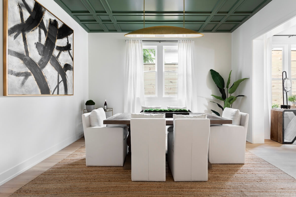 Dining room with a wooden table, six white slipcovered chairs, and a green coffered ceiling, featuring a large abstract black and white painting on the wall.