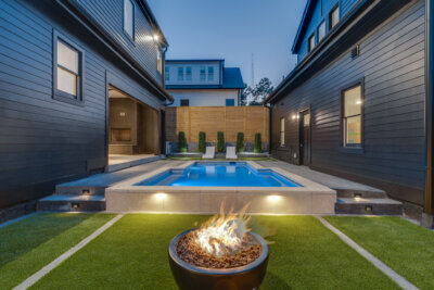 A backyard patio with a lit fire pit in the foreground, a rectangular swimming pool, and two houses with dark siding.