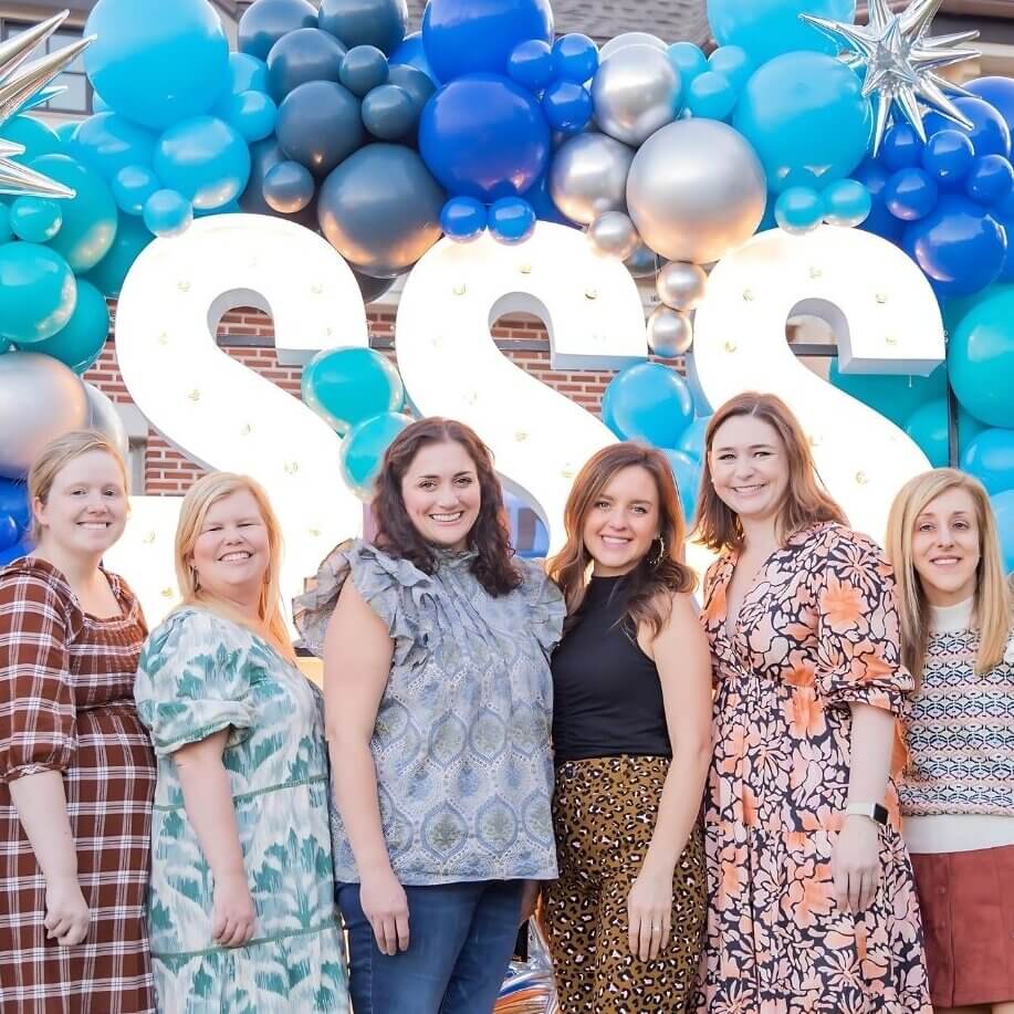 Eight women from the Junior League of Birmingham stand in front of blue balloon decorations and large illuminated letters at an outdoor event.