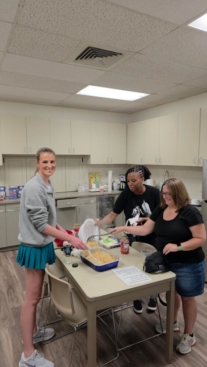 Three women from the Junior League of Birmingham prepare food together in a kitchen with beige cabinets, pasta, and ingredients spread across the table.