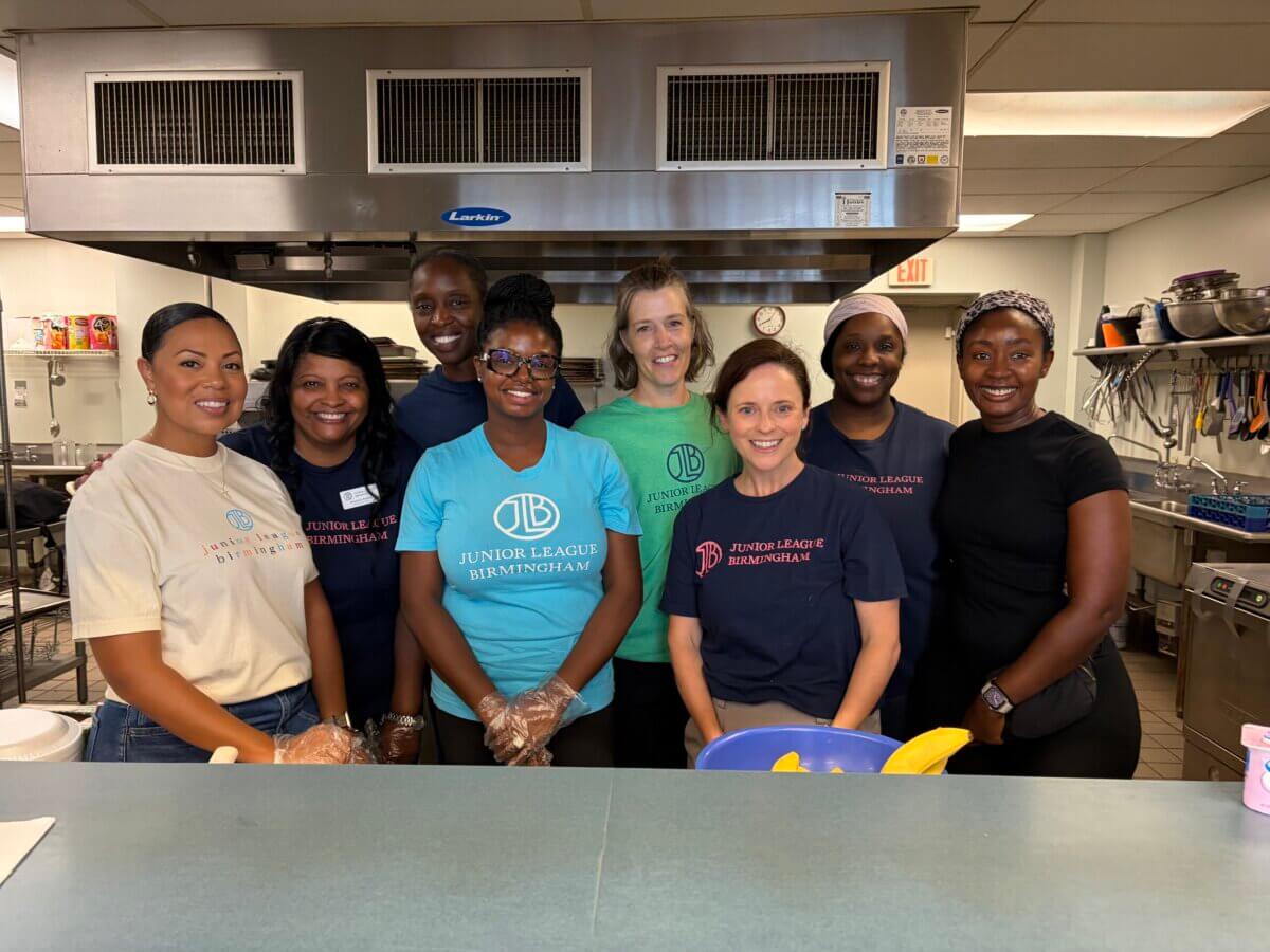 Eight women from the Junior League of Birmingham stand together in a commercial kitchen, smiling at the camera with kitchen equipment visible behind them.