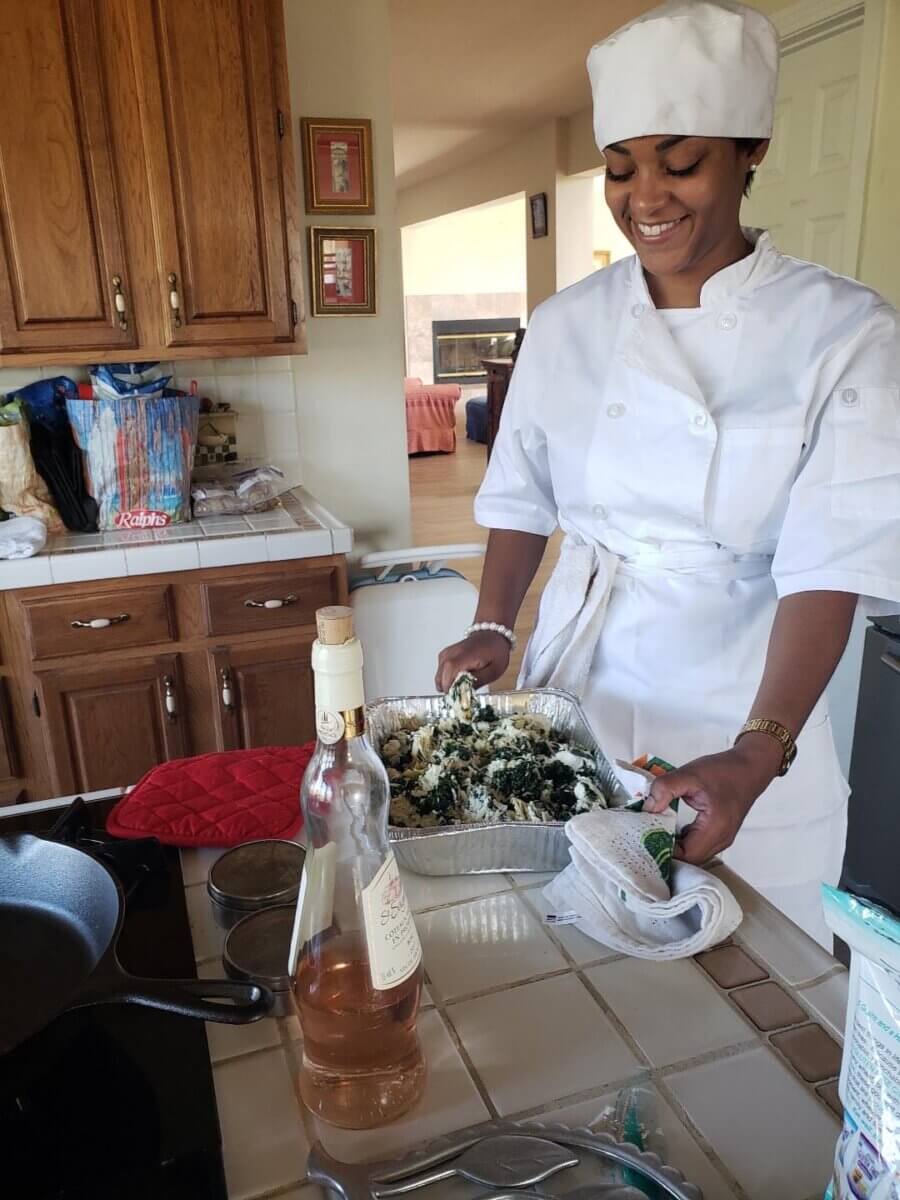 Southern Roots, Japanese Technique: Inside Chef Ashley Jackson's Johin'na - 4 A smiling chef in a white uniform uses a Japanese technique to prepare a metal pan of spinach and cheese pasta in a kitchen.