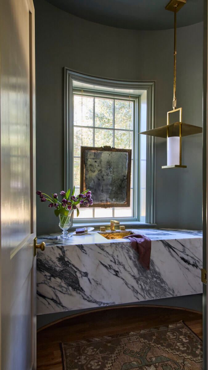 Your Home Should Tell Your Story — and Leigh Misso Wants to Help You Write It - 6 A River Brook Design and Construction bathroom vanity features a marble countertop, a small vase of purple tulips, and a mirror suspended in front of the window.