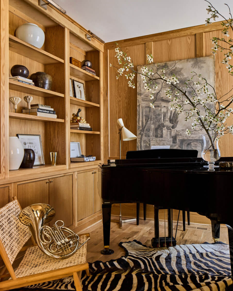 Your Home Should Tell Your Story — and Leigh Misso Wants to Help You Write It - 10 A black grand piano stands before a wall with custom home design shelving and a framed mural, with a French horn on a wicker chair in the foreground.