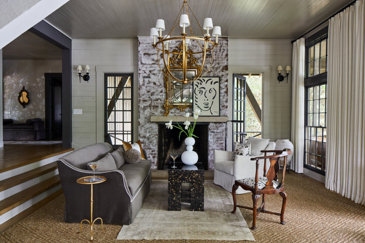 Your Home Should Tell Your Story — and Leigh Misso Wants to Help You Write It - 7 A River Brook Design living room features a brick fireplace, gold chandelier, grey sofa, white armchair, and antique wooden chair arranged around a patterned coffee table.