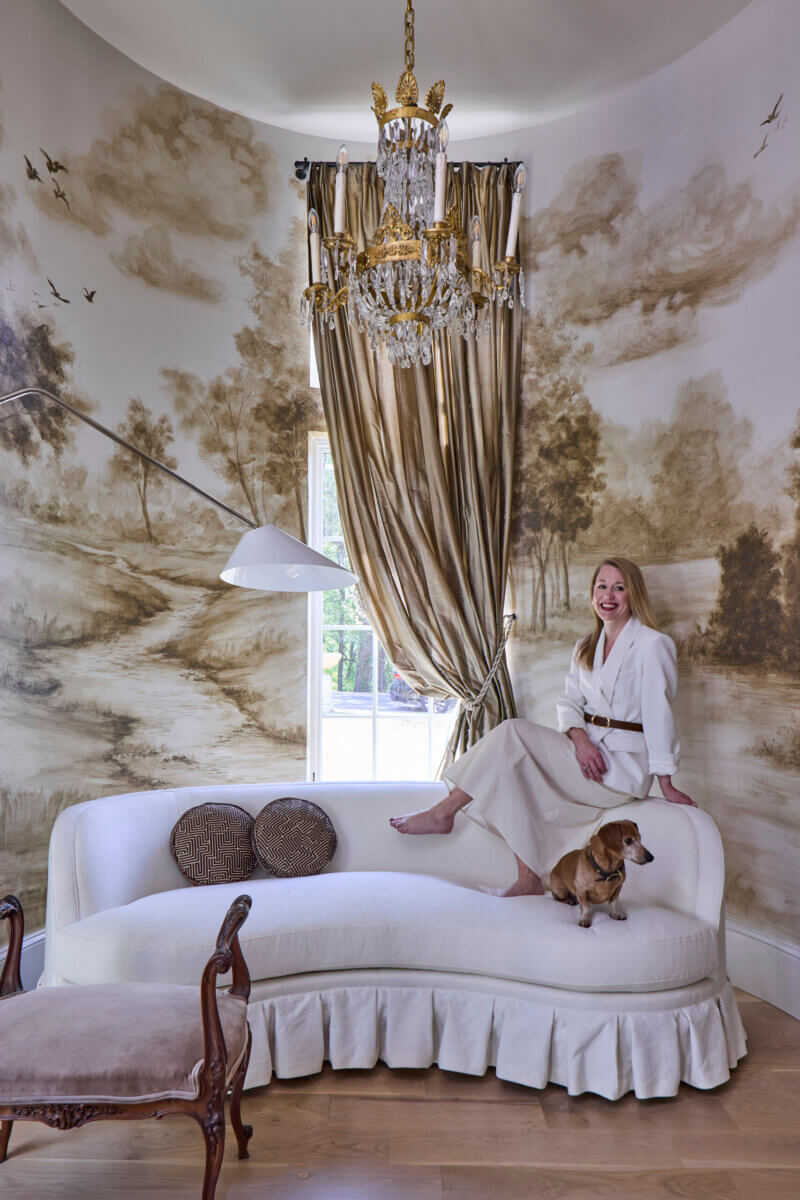 Your Home Should Tell Your Story — and Leigh Misso Wants to Help You Write It - 17 Leigh Misso sits on a curved white sofa with a small dog in a room featuring River Brook Design, a gold chandelier, and mural-painted walls.