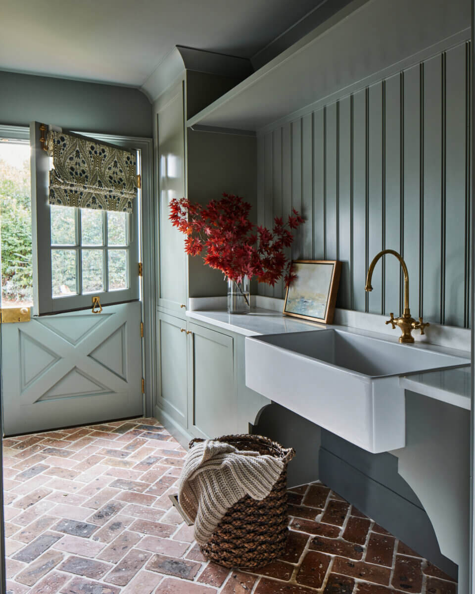 Your Home Should Tell Your Story — and Leigh Misso Wants to Help You Write It - 14 A River Brook Design and Construction mudroom with a white farmhouse sink, brass faucet, and dutch door opening to the outdoors, featuring a basket on a brick floor.