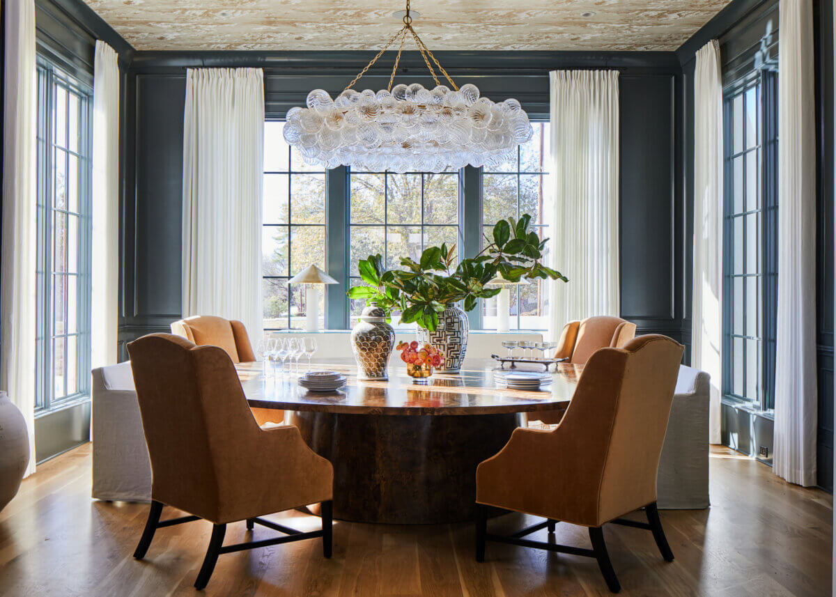 Your Home Should Tell Your Story — and Leigh Misso Wants to Help You Write It - 13 A round wooden dining table surrounded by six upholstered chairs under a glass bubble chandelier in a dark-walled dining room designed by River Brook Design and Construction.