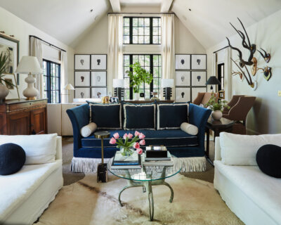 A living room by River Brook Design and Construction features a blue sofa, white armchairs, and a glass-topped coffee table with pink tulips under a vaulted ceiling with large windows.