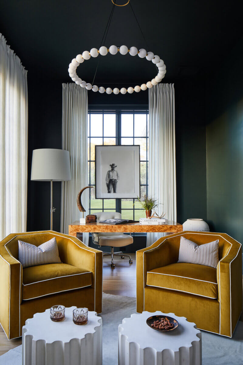 Your Home Should Tell Your Story — and Leigh Misso Wants to Help You Write It - 15 A home office by Leigh Misso featuring two mustard yellow armchairs, two scalloped white side tables, and a wooden desk beneath a round pendant light against dark green walls.