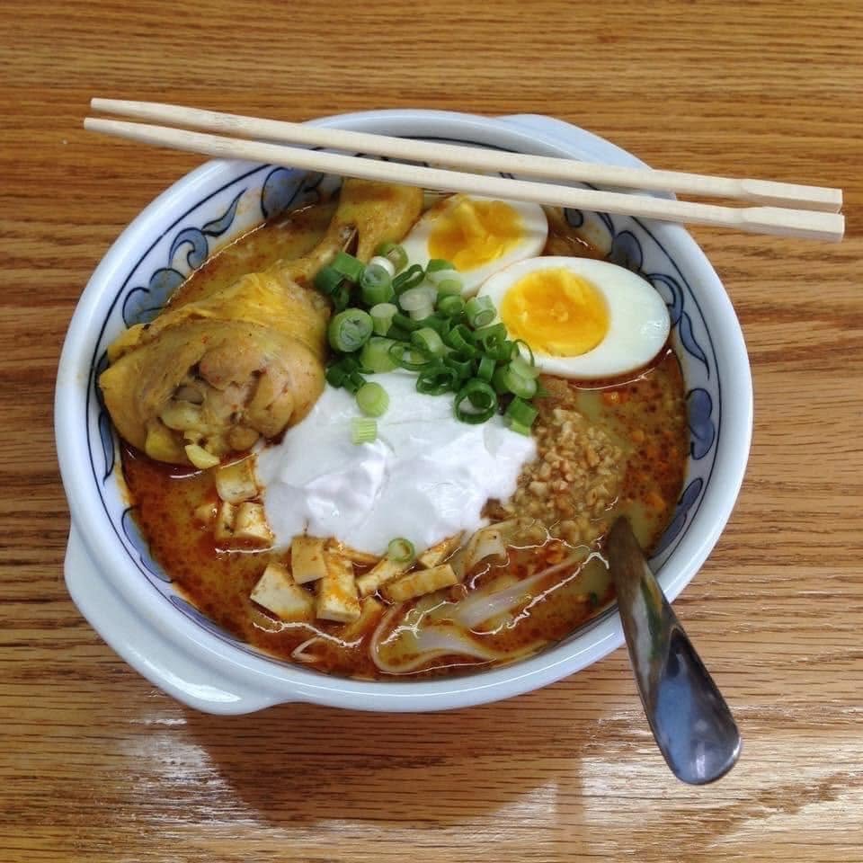 5 New Birmingham Restaurants to Try This Month (Plus An Expansion & A Relocation!) - 3 A bowl of laksa topped with a chicken drumstick, halved soft-boiled egg, tofu cubes, and chopped green onions, with chopsticks resting on the rim.