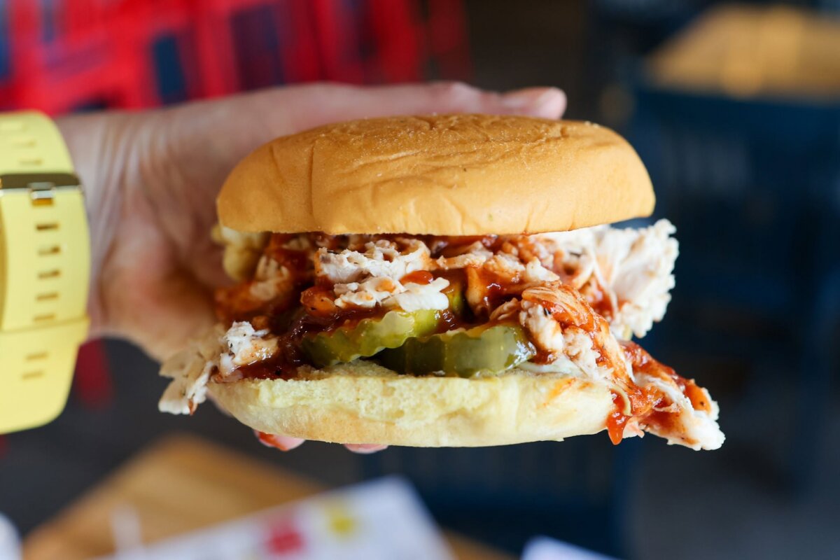 5 New Birmingham Restaurants to Try This Month (Plus An Expansion & A Relocation!) - 6 A hand holds a pulled chicken sandwich with pickles and barbecue sauce on a bun.