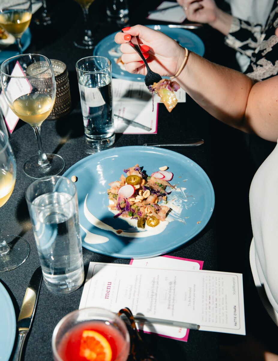 What Happens When You Bring 22 Birmingham Strangers Together and Say "Trust Me"? - 5 A hand holds a forkful of food over a blue plate of salad with radishes and jalapenos, set at a dinner table with wine glasses and a Hattie O’Hara menu for Trust Me Night in Birmingham.