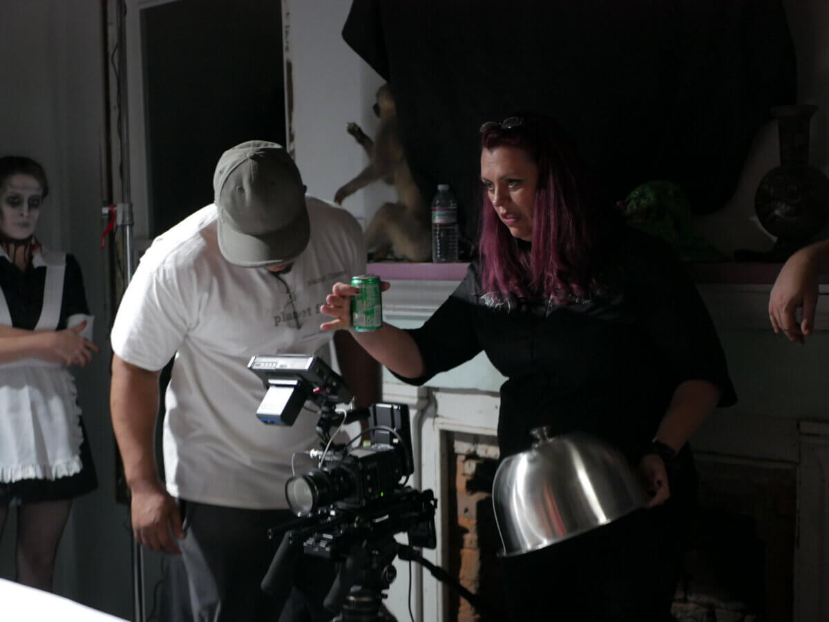 Shannon Ford Thompson holds a green beverage can and a silver serving dome while a camera operator films her; a person in a maid costume stands in the background.