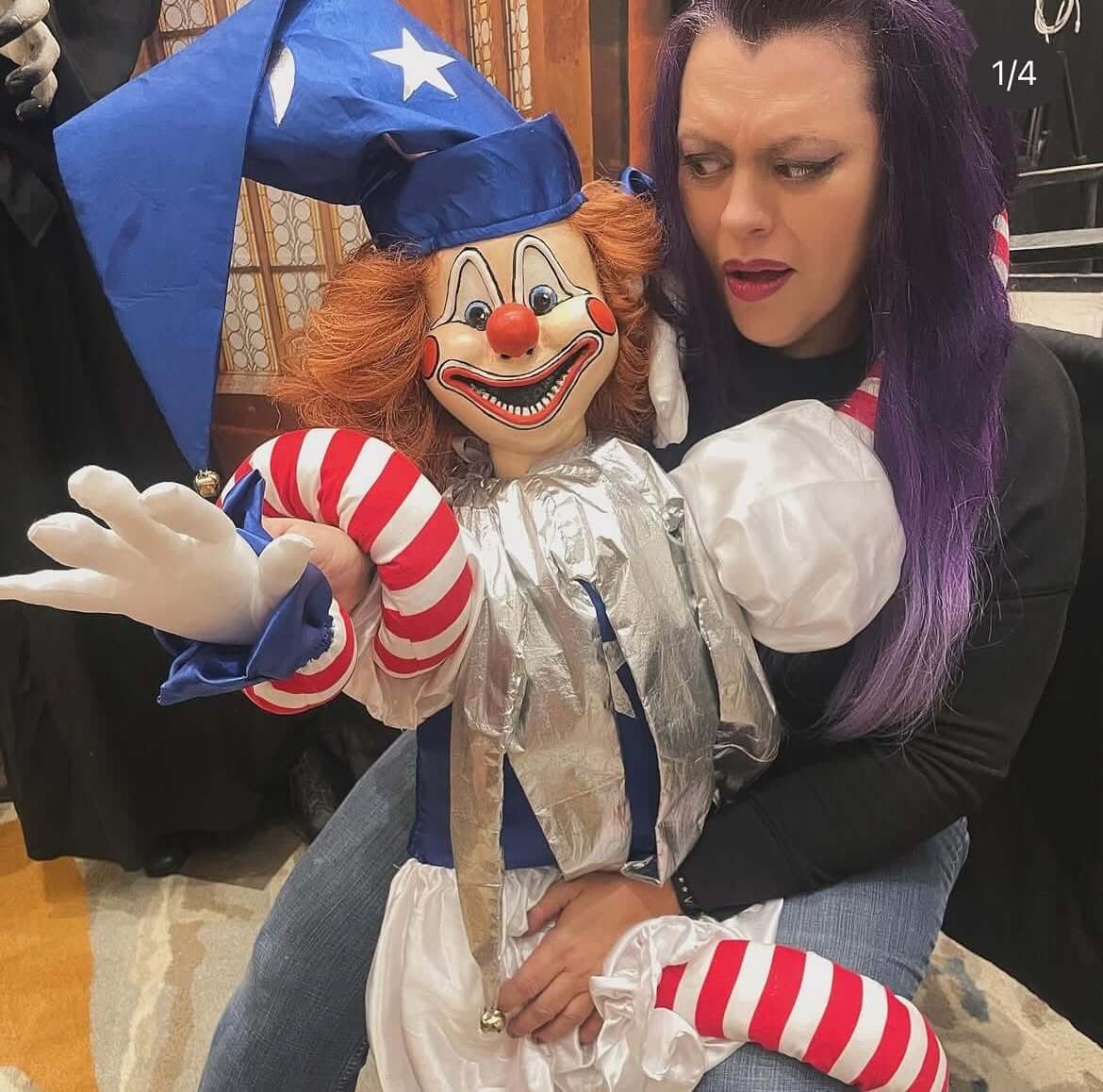 A woman with purple hair looks skeptically at a ventriloquist-style clown doll wearing a blue hat and silver suit.