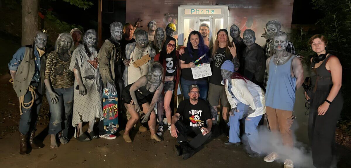 Influencer Shannon Ford and a group of people in zombie makeup pose for a photo outside a prop phone booth, with one person in the center holding a film clapperboard.