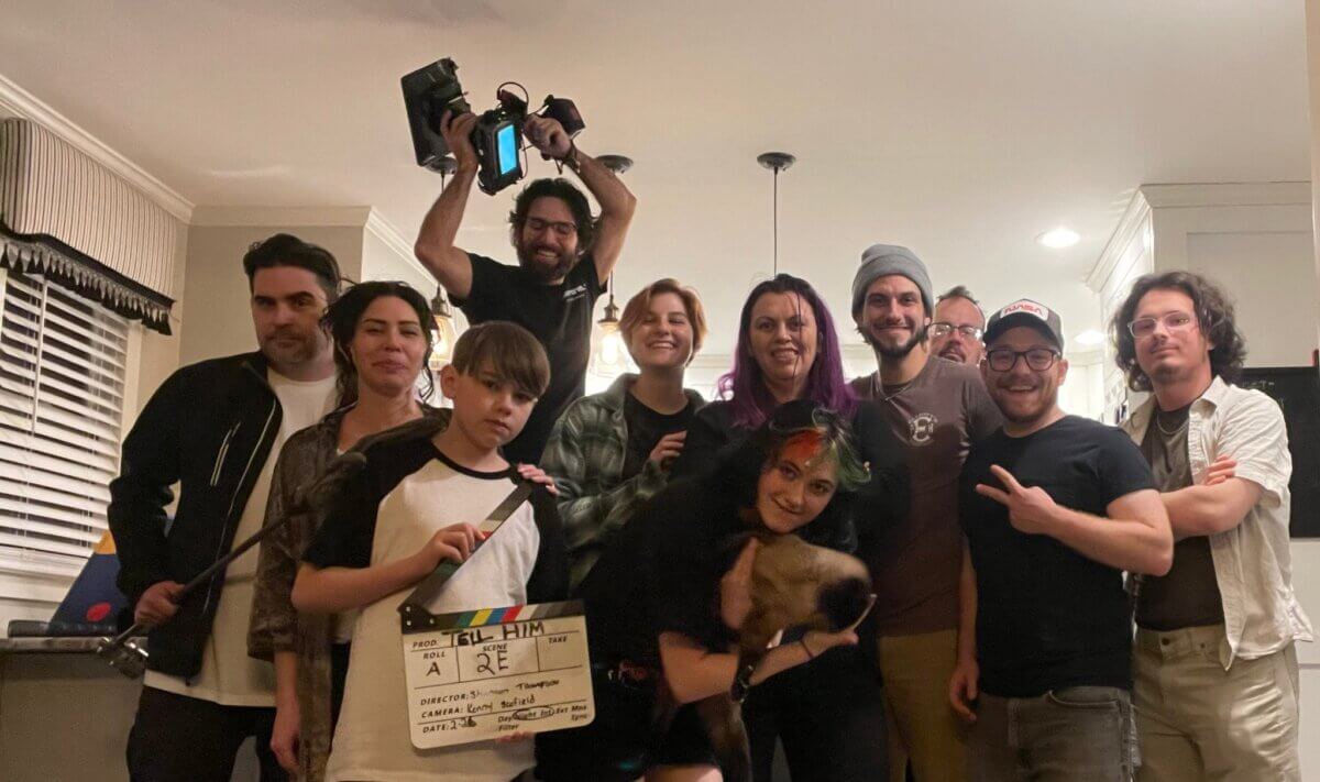 A film crew poses for a group photo indoors, with one person holding a camera above their head and another holding a film slate.