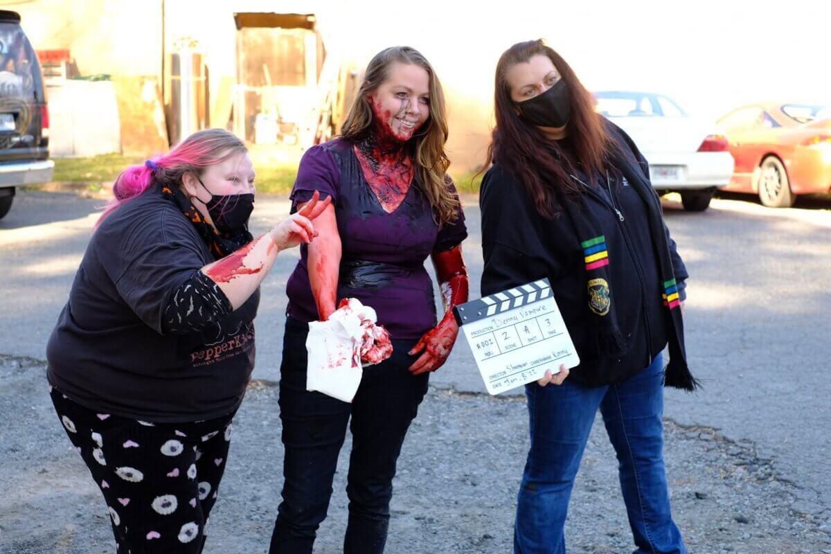 Three women on a film set, one with stage makeup of blood on her face and arms, holding a movie clapperboard.