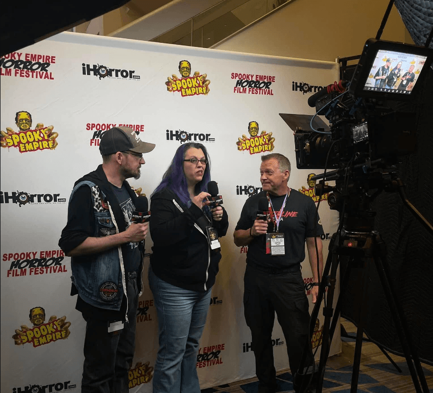 Two men and a woman holding microphones conduct an interview in front of a Spooky Empire Horror Film Festival step-and-repeat banner, while being filmed by a professional camera on a tripod.