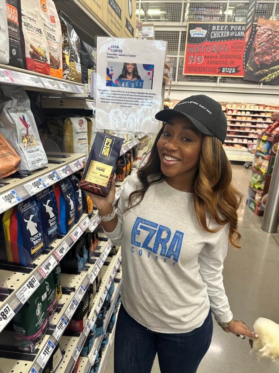 She Started Roasting Coffee on Her Stovetop. Now Target & Amazon Are Calling. - 5 Woman wearing an Ezra Coffee shirt and hat holds a bag of Ezra Coffee in a grocery store aisle, smiling at the camera.