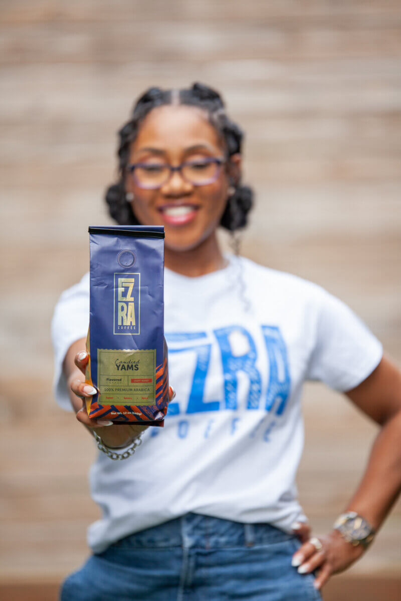 She Started Roasting Coffee on Her Stovetop. Now Target & Amazon Are Calling. - 3 A woman wearing glasses and an Ezra Coffee t-shirt holds a bag of Ezra Coffee toward the camera, smiling.