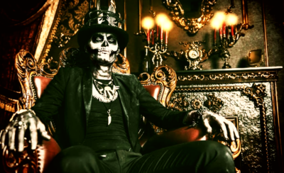 A person in a black suit, top hat, and skull-style face paint sitting on an ornate armchair, ready for a ghost tour.
