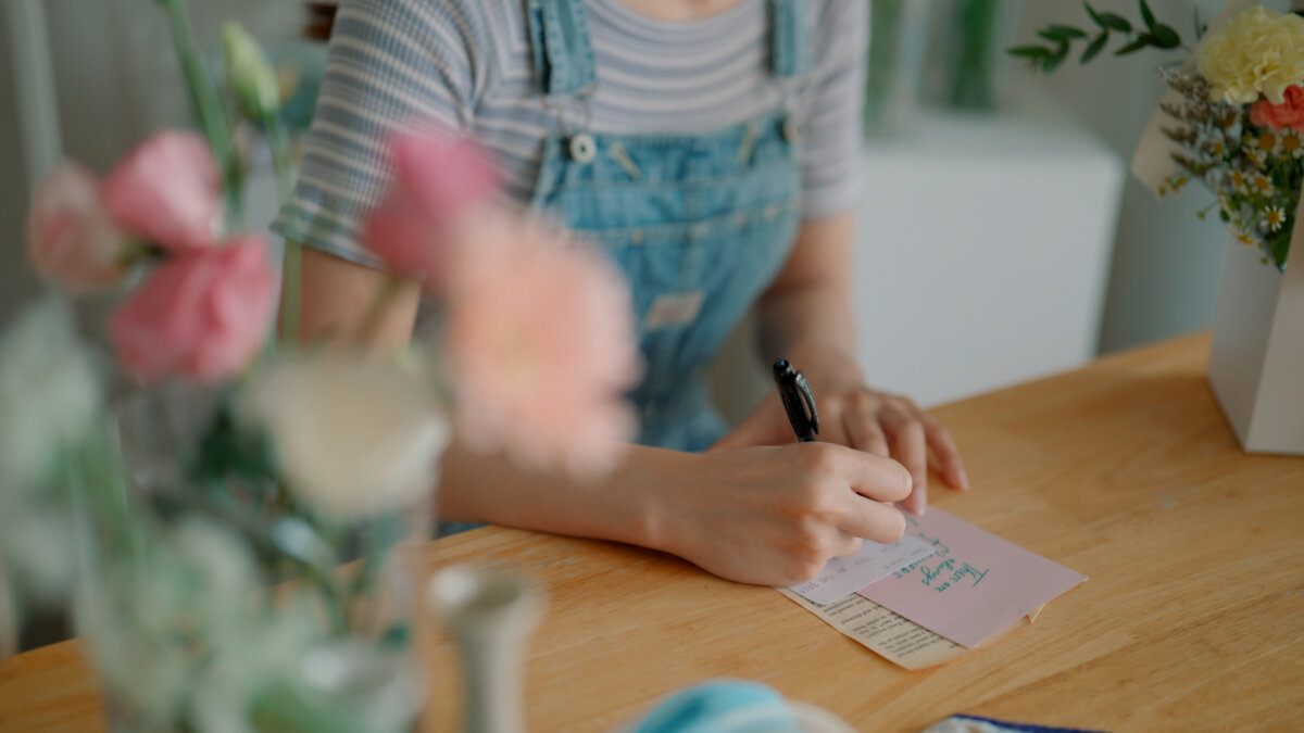 Grammar Guru: Compliment vs. Complement and Stationary vs. Stationery - 3 A person in denim overalls writes on a pink stationery note card at a wooden table surrounded by flowers.