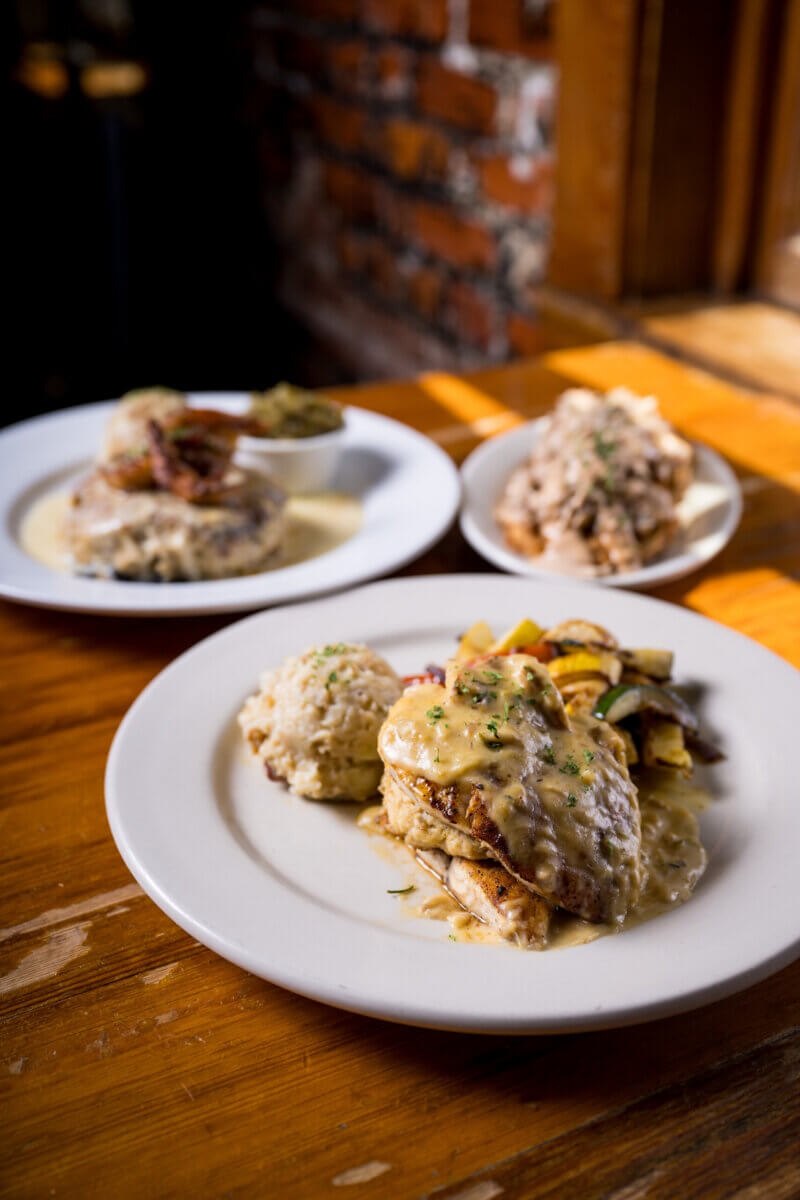 Our Favorite Historic Downtowns in the South to Explore - 7 Three plates of food on a wooden table, featuring grilled chicken in creamy sauce—classic flavors from the South—set against a sunlit brick wall in a charming historic downtown.