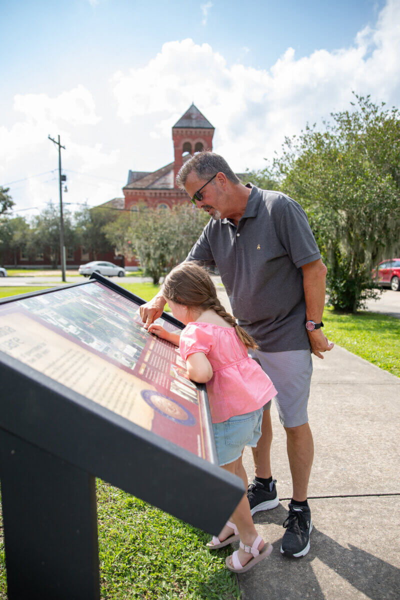Our Favorite Historic Downtowns in the South to Explore - 6 A man and a young girl read an outdoor informational display in the South, with a red brick building and green trees in a historic downtown under a partly cloudy sky.