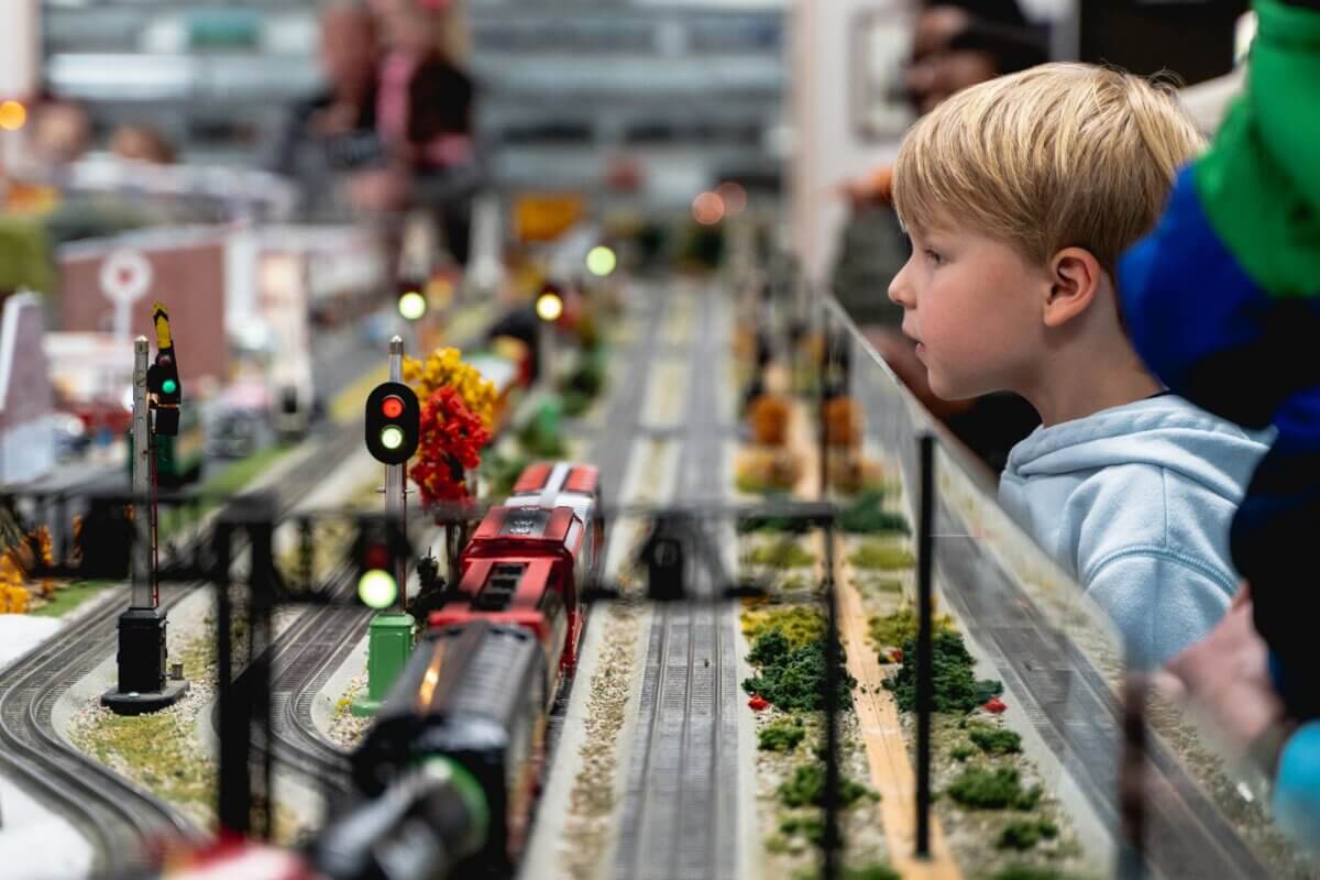 Our Favorite Historic Downtowns in the South to Explore - 5 A young boy closely observes a model train set on display, featuring tracks, miniature trains, and signal lights—reminiscent of railroads that once wove through historic downtowns in the South—while other people linger in the background.