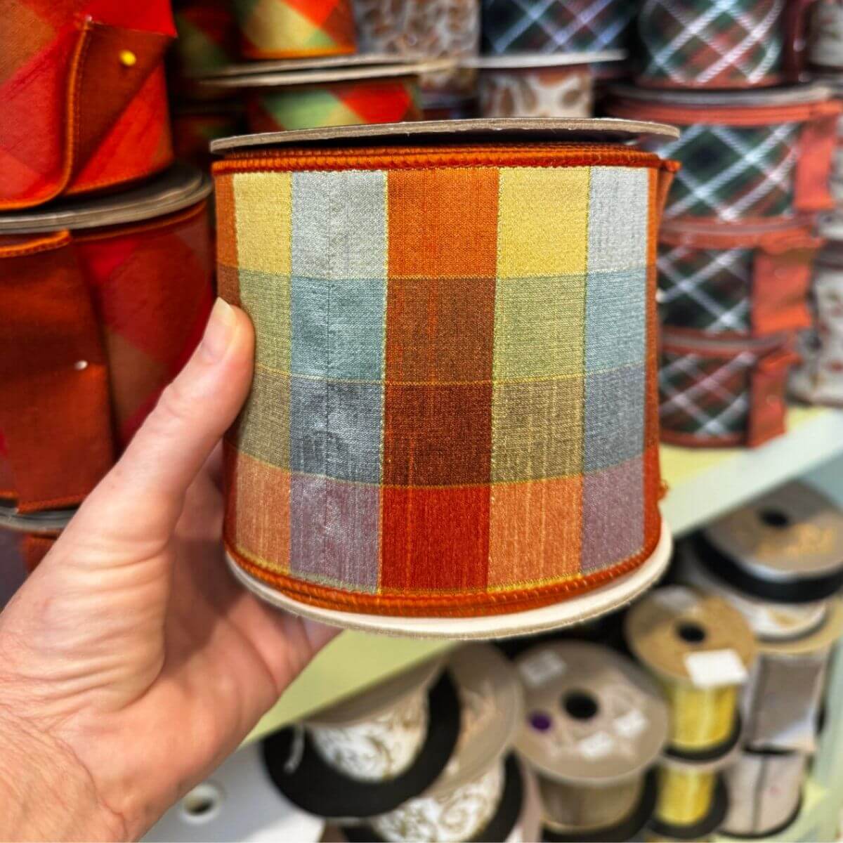 17 Louisville FINDS for October: Fall Fashion, Festive Decor & Game-Day Essentials - 9 A hand holds a roll of plaid ribbon featuring orange, yellow, green, and blue checked patterns—perfect for Louisville finds this October. Other ribbon rolls are stacked in the background.